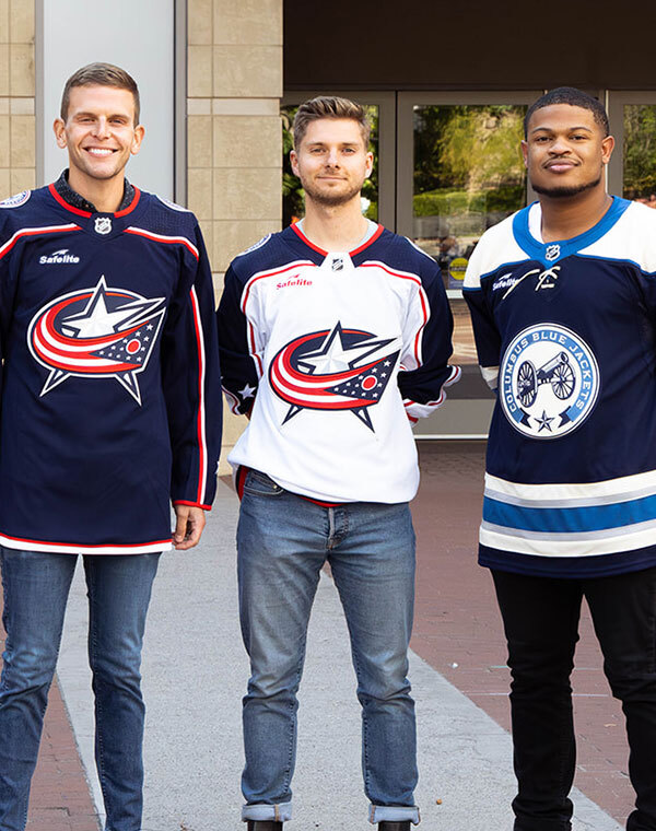 Three men wearing Columbus Blue Jacket Jerseys in front of the arena